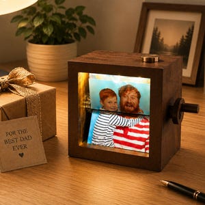 Könnte beinhalten: Ein Holz-Fotowürfel mit Drehmechanismus, der ein Familienfoto zeigt. Eine Geschenkbox mit Schleife und einer Karte mit der Aufschrift "FOR THE BEST DAD EVER" befindet sich neben dem Würfel. Eine Schreibtischlampe und eine Topfpflanze sind im Hintergrund.