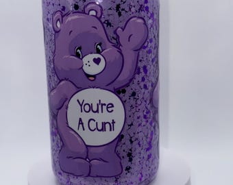 May include: A purple glitter tumbler featuring a cartoon bear with a heart on its belly. The text "You're A Cunt" is printed on the bear's belly. The tumbler has a purple lid and is filled with purple glitter.