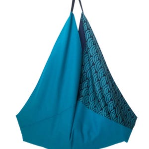 Op de afbeelding: Een turquoisekleurige draagtas met een uniek ontwerp. E&eacute;n kant is effen turquoise, terwijl de andere een turquoise en marineblauw golfpatroon heeft. De tas heeft een donkerblauwe handgreep en een driehoekige vorm.