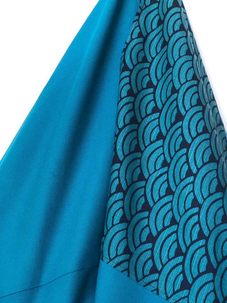 Op de afbeelding: Close-up van een teal stof met een geometrisch patroon. De stof is effen teal van kleur met een donkerder teal patroon op de mouw. Het patroon is een reeks overlappende gebogen vormen.
