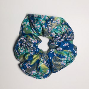 May include: A blue fabric hair scrunchie featuring a repeating pattern of small pink cherry blossoms and gold-colored abstract shapes. The accessory has a gathered, ruffled texture and a circular shape with a hollow center for hair styling.