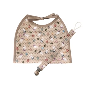 May include: A baby bib and pacifier clip set. The bib is beige with a repeating pattern of white rabbits, flowers, and geometric shapes. The bib has a beige trim and a bow closure. The pacifier clip matches the bib's pattern.