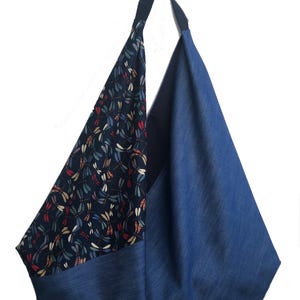 Puede incluir: Un bolso tote de mezclilla azul con una sola correa para el hombro. Un lado presenta una tela azul oscuro estampada con coloridas lib&eacute;lulas. El bolso tiene forma triangular y est&aacute; dise&ntilde;ado para transportar objetos.