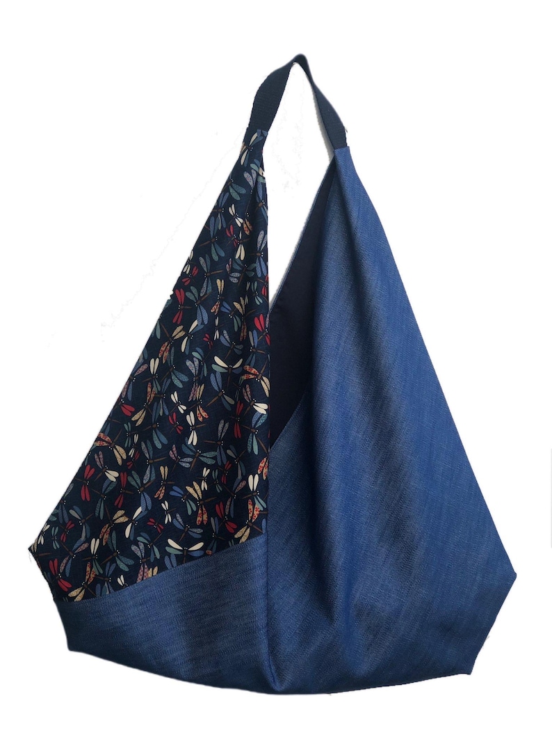 Puede incluir: Un bolso tote de mezclilla azul con una sola correa para el hombro. Un lado presenta una tela azul oscuro estampada con coloridas lib&eacute;lulas. El bolso tiene forma triangular y est&aacute; dise&ntilde;ado para transportar objetos.