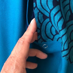 Op de afbeelding: Close-up van een turquoise kledingstuk met een patroonstofdetail. De patroonstof heeft een teal en marineblauw ontwerp. Een hand raakt het kledingstuk aan en onthult een zilveren metalen oogje.
