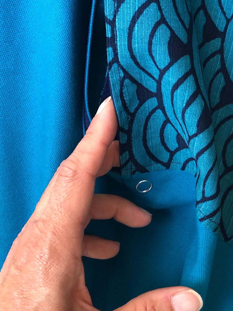 Op de afbeelding: Close-up van een turquoise kledingstuk met een patroonstofdetail. De patroonstof heeft een teal en marineblauw ontwerp. Een hand raakt het kledingstuk aan en onthult een zilveren metalen oogje.