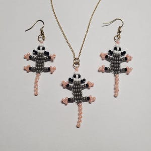 May include: A beaded jewelry set featuring a possum design. The set includes a necklace and a pair of earrings. The possums are gray, black, and white with pink accents. The necklace has a gold chain, and the earrings have gold-colored hooks.