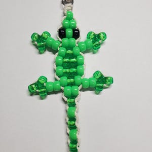 May include: A green beaded lizard keychain with black eyes and a silver clasp. The lizard's body is made of translucent green beads, and the tail is braided with white string. The keychain is a fun accessory.