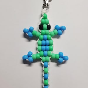 May include: A handmade beaded lizard keychain in shades of green and blue, with black bead eyes. The lizard is attached to a silver-colored metal clasp and white cord. The beaded lizard is a unique accessory.
