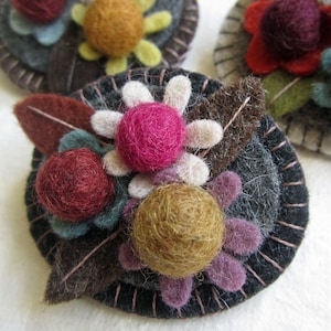 May include: A circular felt brooch featuring layered dark grey fabric with visible pink stitching along the edge. The surface is decorated with three-dimensional needle-felted spheres in magenta, deep red, and mustard yellow, surrounded by felt petals and leaves in various earthy tones.