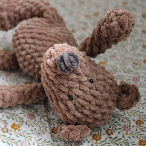 May include: A brown crocheted stuffed animal with a soft, textured yarn finish. The plush toy features a rounded head, small dark eyes, a grey nose, and long limbs. The body is crafted from thick, plush yarn, creating a soft and tactile appearance.