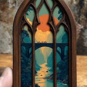May include: A dark brown wooden frame shaped like a pointed gothic arch containing a printed illustration of a stone bridge over a river. The scene depicts a group of small figures walking across the bridge against a large orange sun and blue sky.