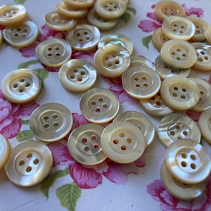 Può includere: Una collezione di bottoni rotondi color crema, ciascuno con quattro fori. I bottoni hanno una lucentezza madreperlacea e sono disposti su una superficie a motivi floreali. Adatti per cucito e progetti di artigianato.