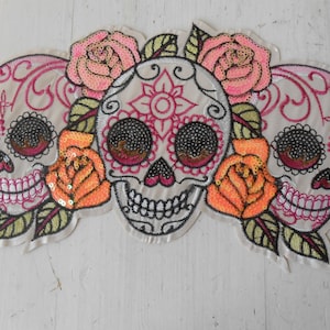 以下が含まれることがあります： ピンクとオレンジのバラと緑の葉が付いた、刺繍された死者の日のスカルデザイン。 このデザインは、お祝いの装飾に最適な、複雑なディテールを備えた3つのシュガースカルを特徴としています。