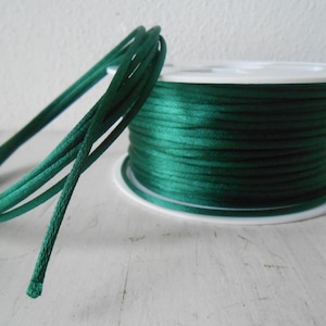 May include: A spool of emerald green cord with a length of cord unwound and looped in the foreground. The cord is a solid, vibrant green color and appears to be made of a smooth, possibly satin-like material. The spool is white.