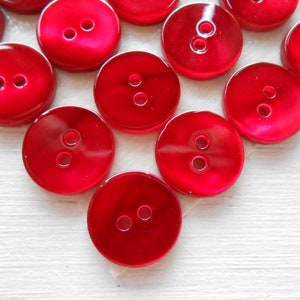 Puede incluir: Un conjunto de botones rojos brillantes y redondos, cada uno con dos agujeros. Los botones están dispuestos cerca, mostrando su color vibrante y textura suave. Ideal para proyectos de costura y manualidades.