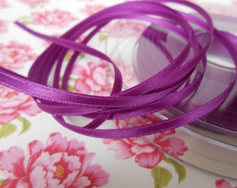Puede incluir: Un carrete de cinta de raso morada vibrante, con un brillo delicado, se muestra sobre un fondo floral. La cinta está dispuesta sueltamente, mostrando su textura y color. Ideal para manualidades y envolver regalos.