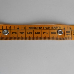 May include: An orange measuring tape with black markings and the text "MISURA PER SARTI". The tape has metal eyelets and measures up to 12 inches. Ideal for sewing and tailoring projects.