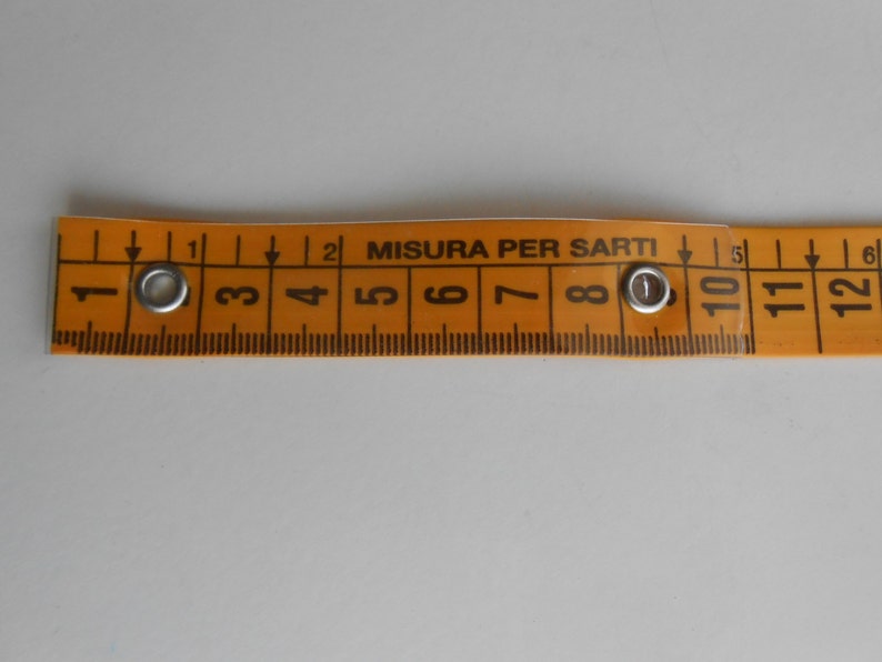 May include: An orange measuring tape with black markings and the text "MISURA PER SARTI". The tape has metal eyelets and measures up to 12 inches. Ideal for sewing and tailoring projects.
