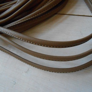 Puede incluir: Primer plano de un cordón de cuero marrón con un borde cosido. El cordón está doblado en forma de U y está sobre una superficie de madera clara.