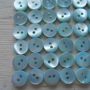 Puede incluir: Colección de botones redondos, azul claro, con dos o tres agujeros cada uno. Los botones tienen un brillo nacarado y están dispuestos en filas ordenadas sobre una superficie clara. Adecuados para costura y manualidades.