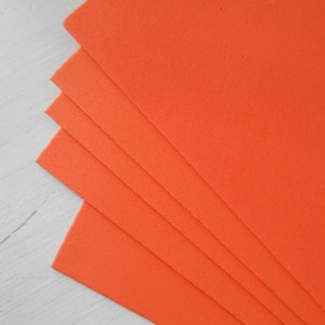 May include: A stack of bright orange foam sheets, fanned out to show their edges. The sheets are a solid, vibrant color, and appear to be made of a porous material. The background is a light, textured surface.