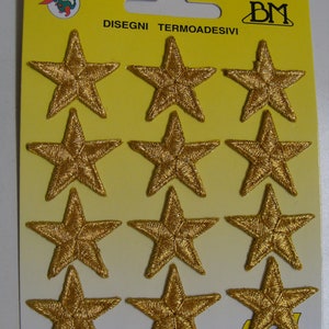 Pode incluir: Uma folha com 12 estrelas douradas para passar a ferro. A folha é amarela com o texto "DISEGNI TERMOADESIVI" e "BM" em fonte preta.