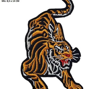 May include: Embroidered iron-on patch featuring a detailed tiger design. The tiger is depicted in a crouching position with its mouth open, showing sharp teeth. The tiger is outlined in black and has orange and white stripes. The patch measures 8.5 by 14 centimeters.