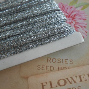 Peut inclure: Une bobine de cordon pailleté argenté, enroulée sur une bobine en carton blanc. La bobine est posée sur un sachet de graines vintage avec le texte "Rosies Seed House Flower"