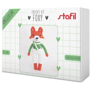 May include: A crochet kit box featuring a handmade fox amigurumi. The fox is orange, white, and green, with a scarf and overalls. The box reads "CROCHET KIT FOXY" and "IN RECYCLED COTTON". The fox is approximately 9.8 inches tall.