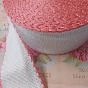 May include: A roll of pink and white trim with a scalloped edge. The trim is made of a woven fabric and has a subtle texture.
