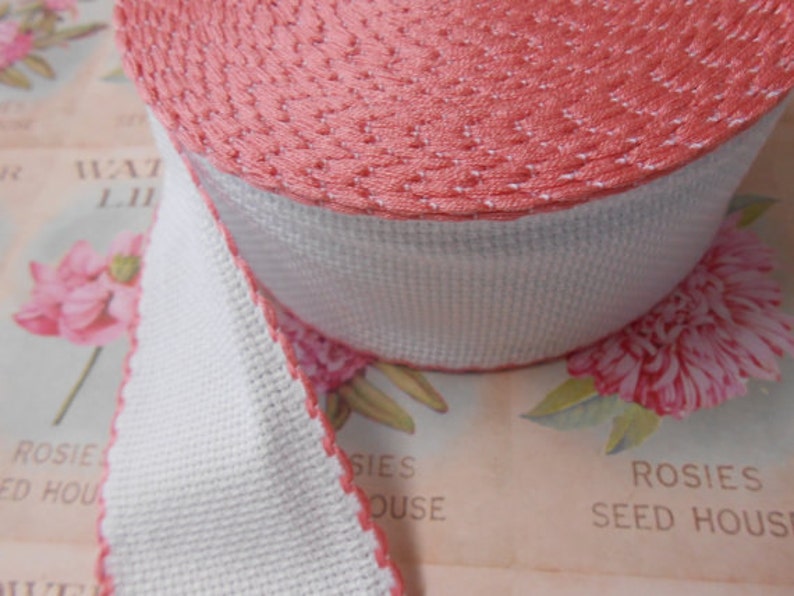 May include: A roll of pink and white trim with a scalloped edge. The trim is made of a woven fabric and has a subtle texture.