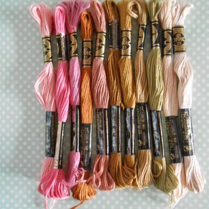 May include: A selection of embroidery threads in shades of pink, orange, gold, and cream. The threads are bundled with black and gold labels, arranged on a pale blue background with white polka dots. Suitable for embroidery and cross-stitch projects.