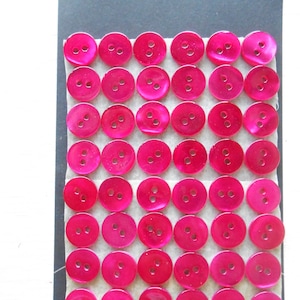 Può includere: Una collezione di bottoni rotondi rosa acceso con tre fori ciascuno, disposti in file su una scheda nera. La scheda ha il testo "VERA MADREPERLA" in alto. I bottoni sono di materiale lucido.