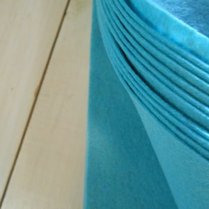 Puede incluir: Primer plano de un trozo de tela de fieltro azul turquesa doblado. La tela está doblada en una serie de pliegues, creando un efecto texturizado y en capas.