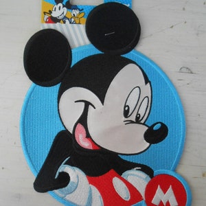 Könnte beinhalten: Ein blauer und weißer gestickter Aufnäher mit Mickey Mouse und einem roten "M" in der unteren rechten Ecke. Der Aufnäher ist rund und hat einen schwarzen Umriss.