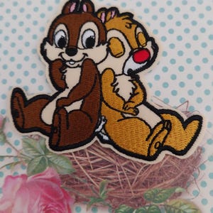 May include: Embroidered patch featuring two cartoon chipmunks, one brown and one tan, sitting back-to-back. The chipmunks have black outlines and are smiling. The patch is on a light beige background with a floral and polka dot design.