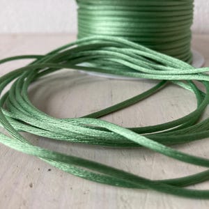 Può includere: Una bobina di cordoncino di raso verde chiaro con una parte del cordoncino srotolata in primo piano. Il cordoncino è di un colore verde tenue e uniforme. Lo sfondo è di colore chiaro e neutro.