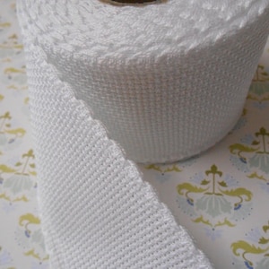 May include: White cotton webbing ribbon, rolled up on a white background. The ribbon is woven with a textured pattern.