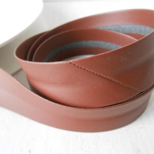 May include: A rolled-up, brown leather strip with a stitched seam. The leather is a warm, reddish-brown color, and the edges are neatly finished. The strip appears to be flexible and durable, suitable for various crafting projects.