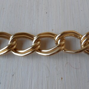Op de afbeelding: Close-up van een goudkleurige ketting met grote, ovale schakels. De ketting heeft een glanzende, metalen afwerking en lijkt van metaal te zijn gemaakt. De schakels zijn in elkaar verweven, waardoor een doorlopend patroon ontstaat. De ketting wordt waarschijnlijk gebruikt voor sieraden of modeaccessoires.
