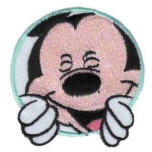 May include: A round, embroidered patch featuring a cartoon character with a pink face, black eyes, and a black nose. The character is wearing a white shirt with a pink bow tie and has white gloves. The patch has a light blue border.