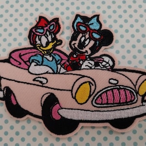 May include: Embroidered patch showing Daisy Duck and Minnie Mouse in a pink convertible car. Both characters wear sunglasses. The patch has a light pink background and black outlines.