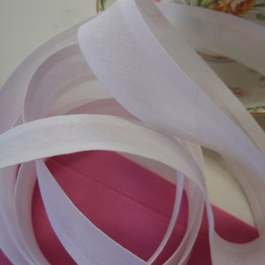 Puede incluir: Cinta de bies de algodón blanco, un tipo de adorno de tela utilizado para terminar costuras y bordes, se muestra en una imagen de primer plano. La cinta está doblada y enrollada, mostrando su textura suave y su color blanco.