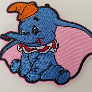 以下が含まれることがあります： 青い象のダンボの刺繍パッチ。オレンジ色の帽子とピンクの耳をしています。パッチには白、赤、青の襟があり、黒で縁取られています。衣類やアクセサリーに加えるのに最適です。