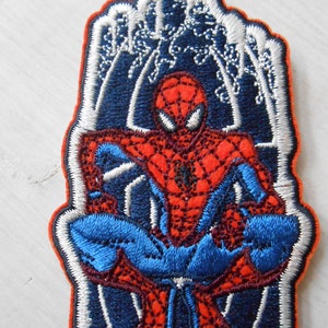 May include: Embroidered iron-on patch featuring a red and blue Spiderman character in a sitting pose. The background is a dark blue with white outlines of a web.