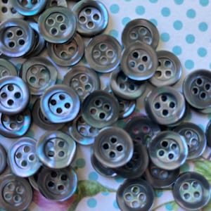 以下が含まれることがあります： 4つの穴のあるグレーの真珠貝ボタンのコレクション。ボタンはランダムなパターンで配置されており、わずかに虹色の輝きがあります。