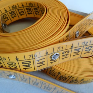 May include: A yellow measuring tape with black numbers and markings. The tape is coiled and partially unrolled, showing measurements in inches and centimeters. The tape is made of a flexible material and is used for sewing and crafting.