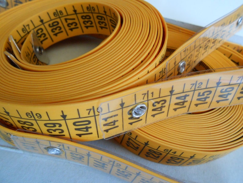 May include: A yellow measuring tape with black numbers and markings. The tape is coiled and partially unrolled, showing measurements in inches and centimeters. The tape is made of a flexible material and is used for sewing and crafting.