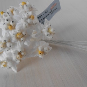 May include: A bunch of white felt edelweiss flowers with yellow centres and black accents. The flowers are attached to thin wire stems. The label reads "351 - 62 Edelweiss 1302 5cm".